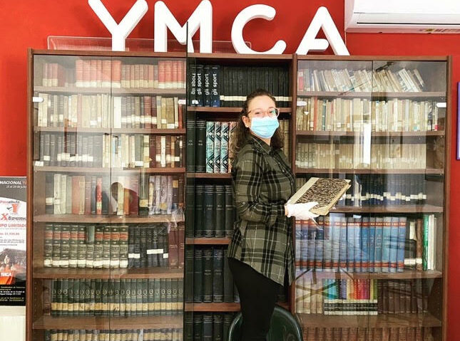 Francesca Natalone, wearing a mask and gloves, in front of a book shelve